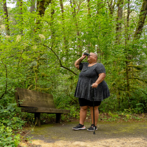 A Black non-binary hiker looks through binoculars in a forest.