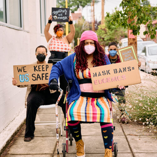 Masked disabled people of color holding protest signs.