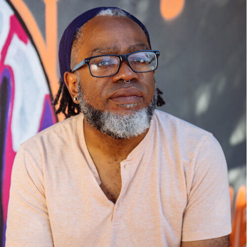A bearded Black man with glasses gazes at the camera neutrally, wearing a peach shirt, septum piercing, small ear gauges, and navy bandana.