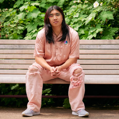 An Asian non-binary person with long hair looks neutrally at the camera while sitting casually on a bench in front of blooming flowers.