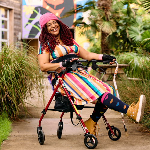 A Black person laughs while sitting back in their rollator and holding a cane.