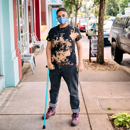 A masked Latinx person stands on a sidewalk with their cane.