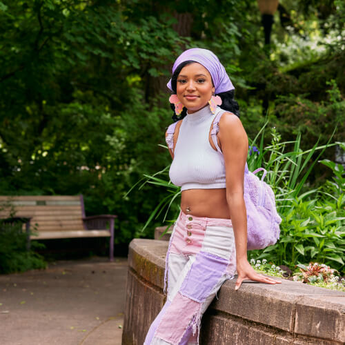 A Black woman with a lavender head scarf, pastel earrings, and white crop tank smiles and stands in the middle of a park.