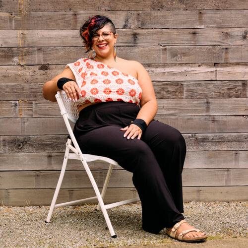 A Native and Latinx woman sits outdoors in a chair and smiles at the camera.