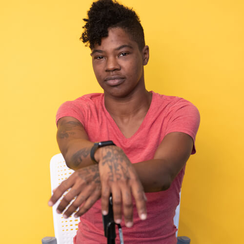 A Black non-binary person sits in front of a yellow backdrop with their arms crossed and resting on their cane.