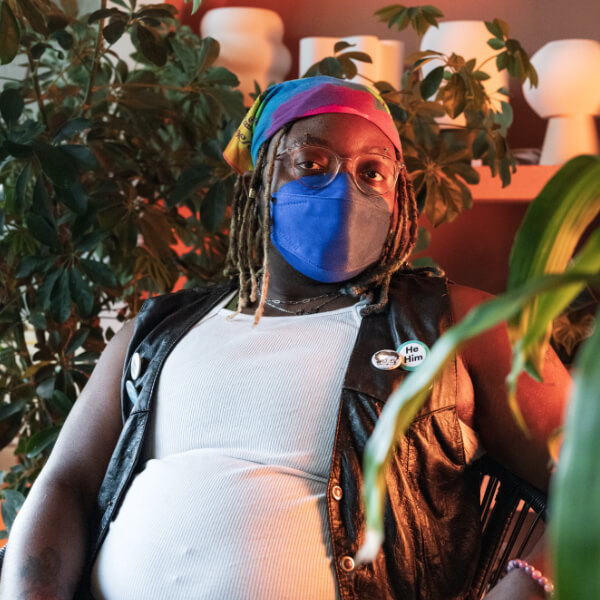 A Black transmasculine person with a face mask and rainbow bandana sits surrounded by indoor plants.