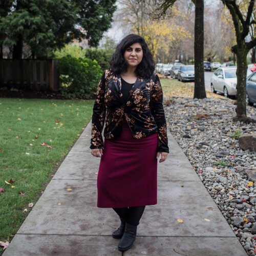 A Latinx woman smiles while standing on a sidewalk on a cloudy day.